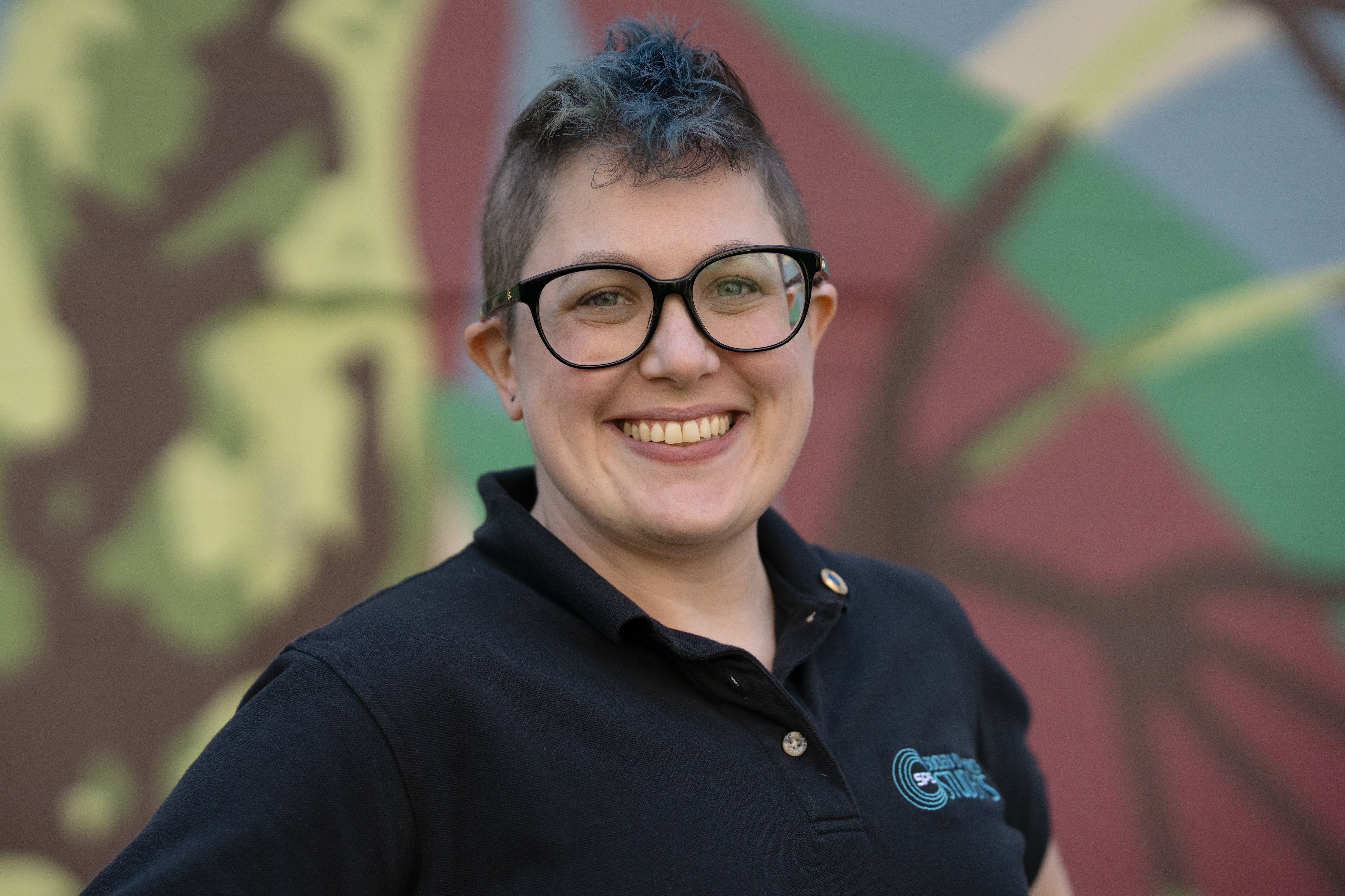 The author, a light-skinned feminine person with a blue faux hawk and glasses wearing a shirt with the logo of the Society of Physics Students